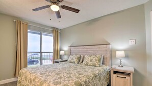 Un dormitorio con una cama grande, un ventilador de techo, una mesita de noche con lámpara y vistas al exterior a través de una ventana.