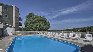 A swimming pool with a metal safety railing, surrounded by white lounge chairs and a building with balconies.