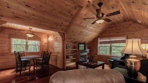 A cozy cabin interior with wooden walls, a dining area, a living room with a sofa and a lamp, and a ceiling fan.