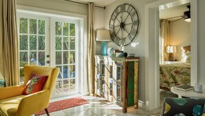 Une pièce avec un fauteuil jaune, une grande horloge murale, une commode rustique et un lit recouvert d’une literie à motifs floraux.