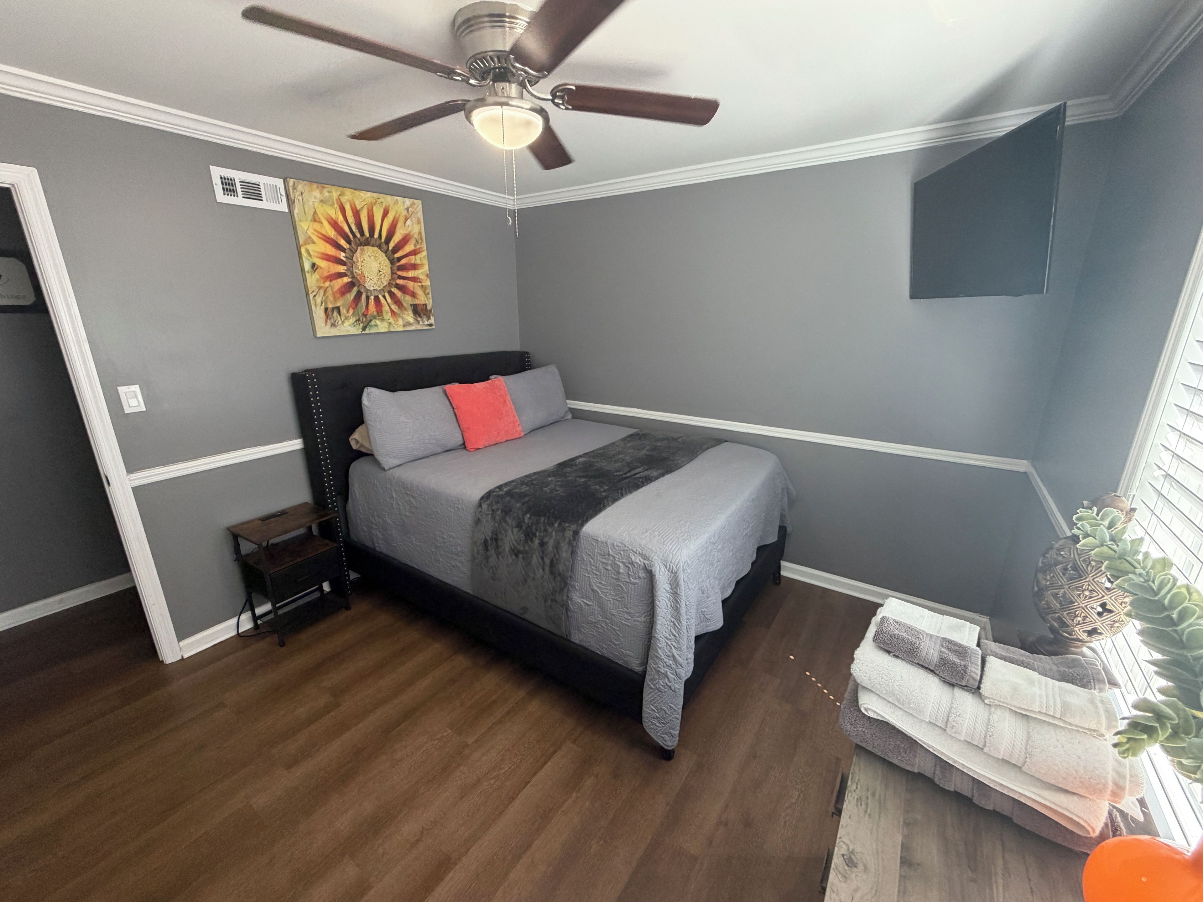 A bedroom with a bed, a ceiling fan, a wall-mounted artwork, a bedside table, and a small plant.