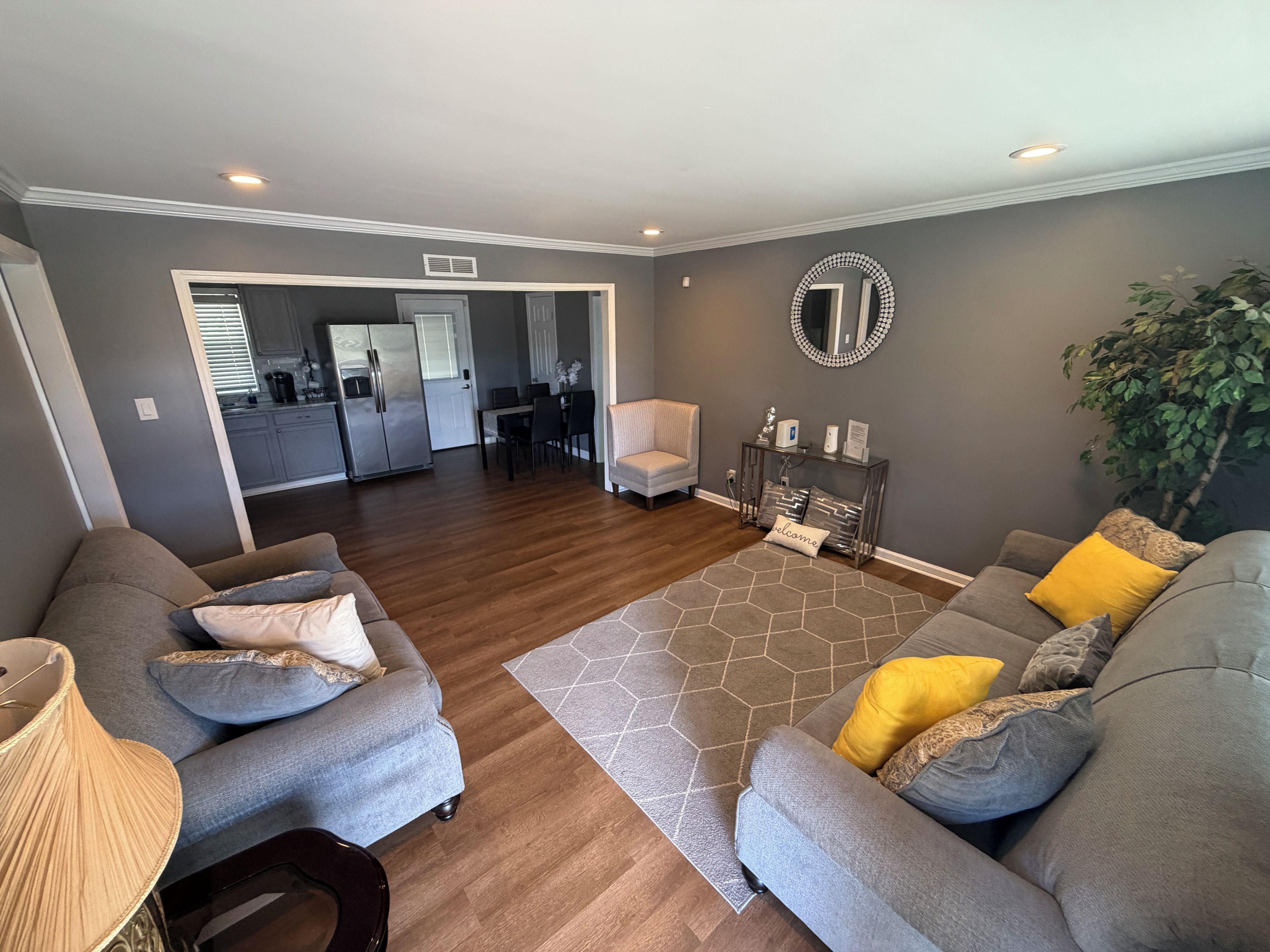 A modern living room with a grey sofa set, a coffee table, and a console table with decorative items. A dining area is visible in the background.