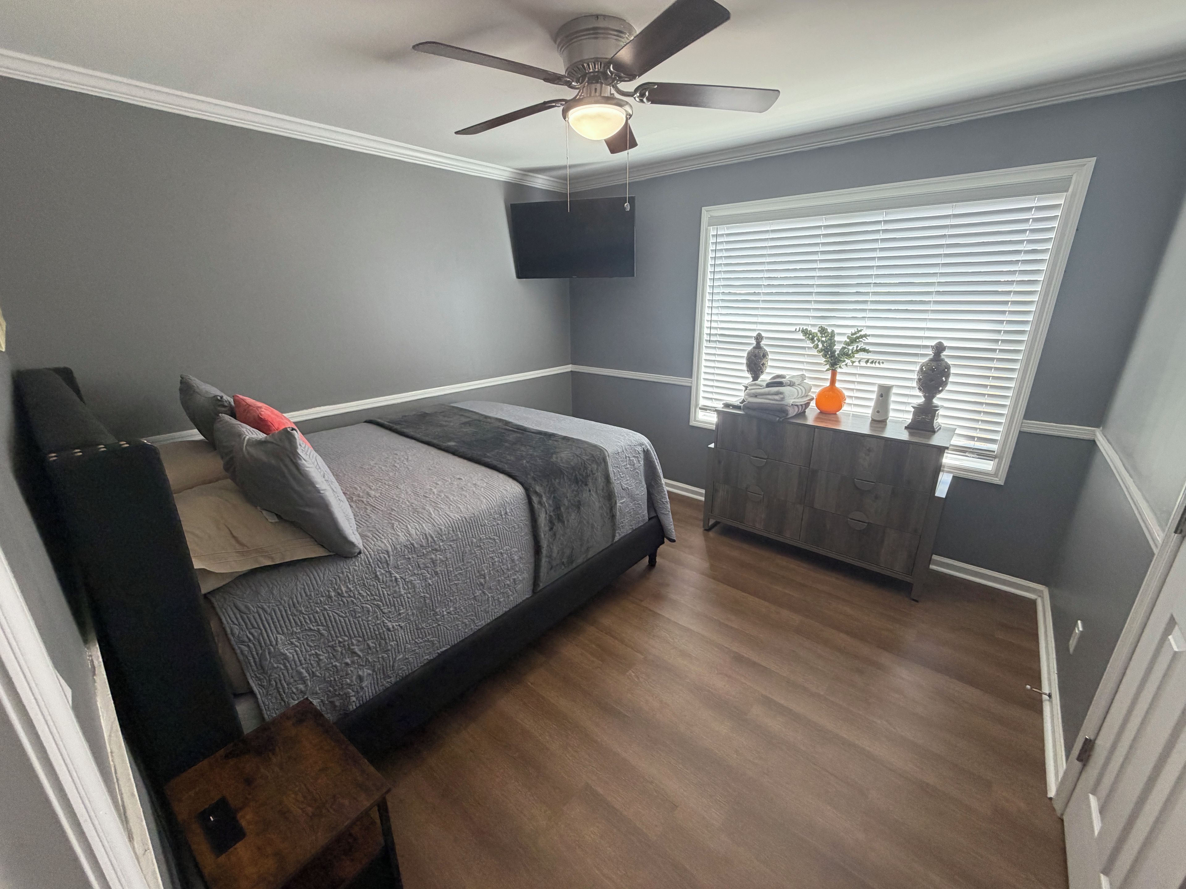 Une chambre à coucher comprenant un lit, une commode, un ventilateur de plafond et une fenêtre avec des stores.