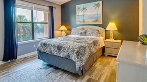 A bedroom with a bed, two bedside tables with lamps, a nightstand, a window with curtains, and a painting of a beach scene. - Walk to Beach: PCB Condo w/ Oceanfront Pool Access (Panama City Beach)