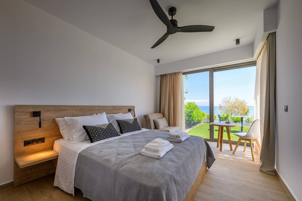 A modern hotel room with a large bed, a ceiling fan, a balcony with a table and chairs, and a view of the sea.