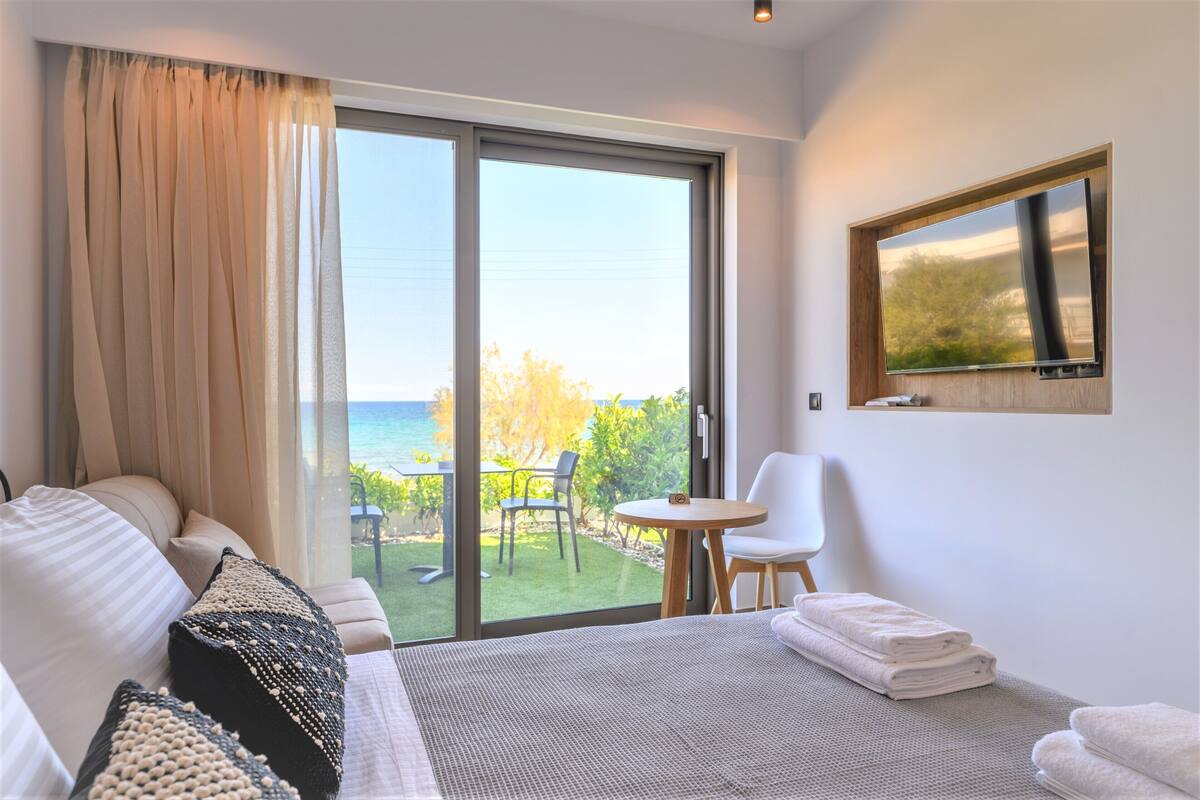 A bedroom with a bed, a small table, a chair, and a view of the ocean through a sliding door.