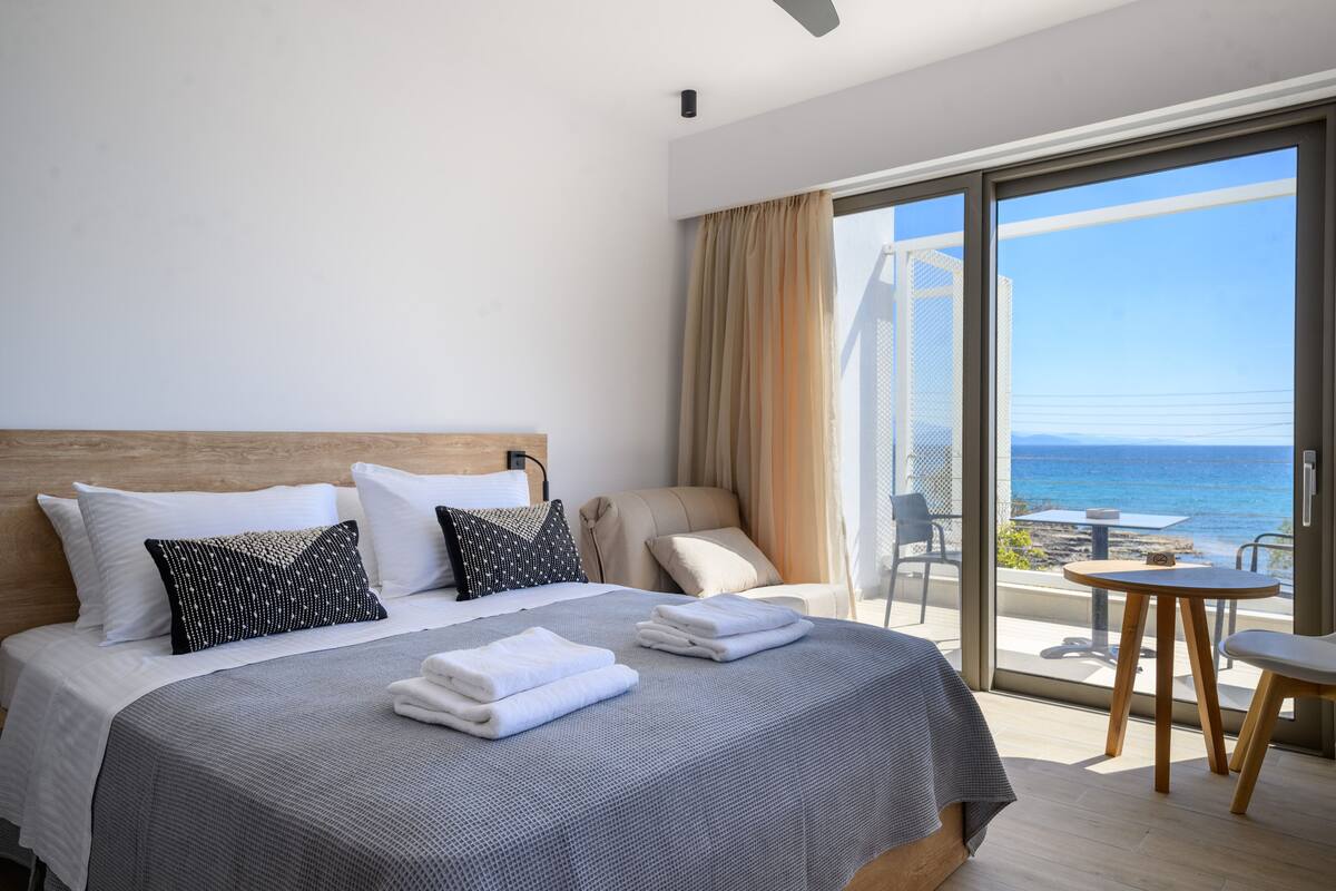 A hotel room with a large bed, a view of the sea, and a balcony with a table and chair.
