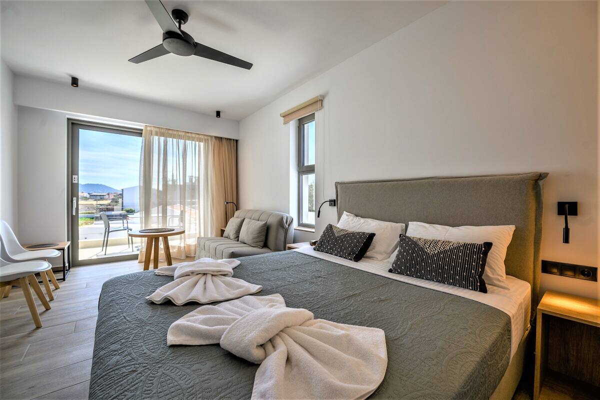 A hotel room with a large bed, a ceiling fan, a balcony with a view, and a seating area with chairs.