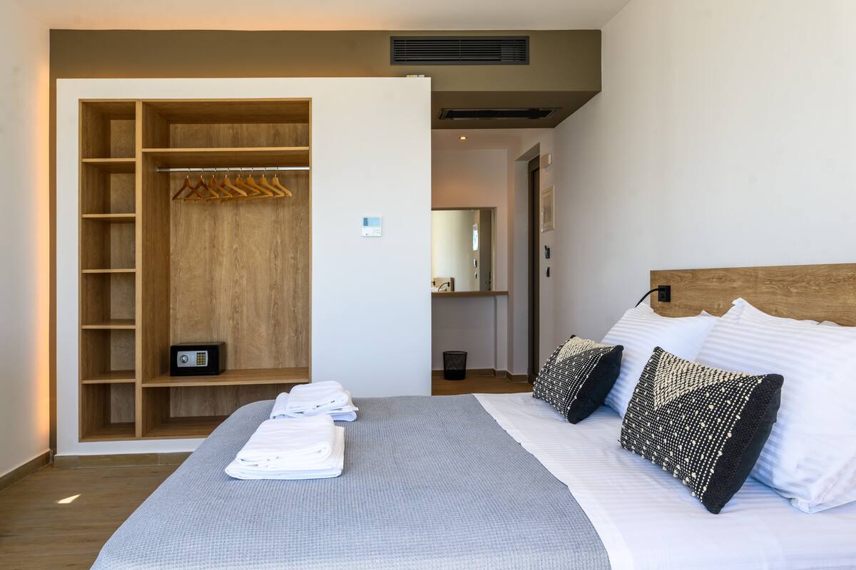 A neatly made bed with white linens and patterned pillows, a wooden wardrobe with hangers, and a small safe on the lower shelf.