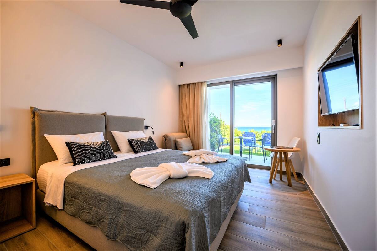 A modern hotel room with a large bed, a ceiling fan, a TV mounted on the wall, and a balcony with a table and chairs.