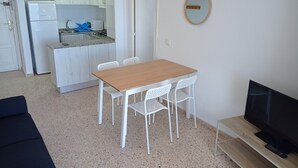 A compact living space with a kitchenette, a dining area with a wooden table and white chairs, and a TV on a wooden stand. - Infante E2-9 Solo Familias! (Playa de Gandía)