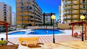 A swimming pool area with a bench, a lifeguard tower, and a view of multi-story buildings in the background. - Infante E2-9 Solo Familias! (Playa de Gandía)