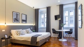Una habitación de hotel moderna con una cama, una mesita, dos sillas, un televisor y ventanas amplias.