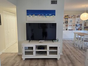 Uma sala de estar com televisão de tela plana em um rack branco, um quadro acima da TV, e uma área de jantar com mesa e cadeiras.