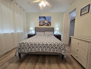 Quarto com cama, abajures, um quadro na parede, cômoda e porta que dá para outro cômodo.