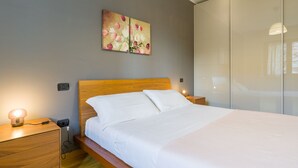 A modern bedroom with a wooden headboard, a bed with white linens, a bedside table with a lamp, and a wall-mounted painting. - Le Volpi in città (Verona)