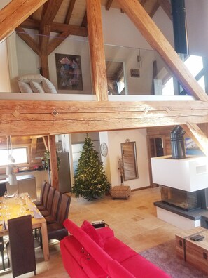 En rummelig loftrum med synlige træbjælker, et spiseområde med et bord dækket til fire personer, en rød sofa og et juletræ.