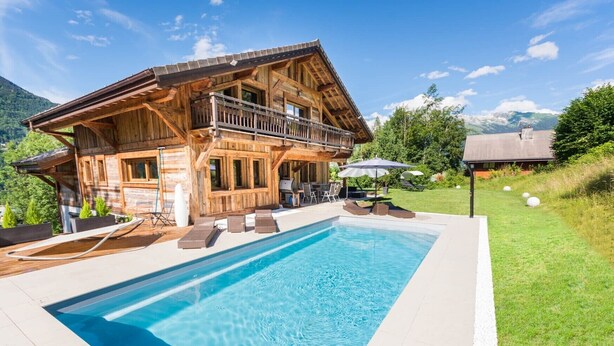 Una cabaña de madera con piscina, mobiliario exterior y montañas al fondo.