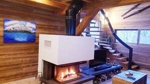 Interior de una cabaña de madera con chimenea, un cuadro de una montaña y una escalera.
