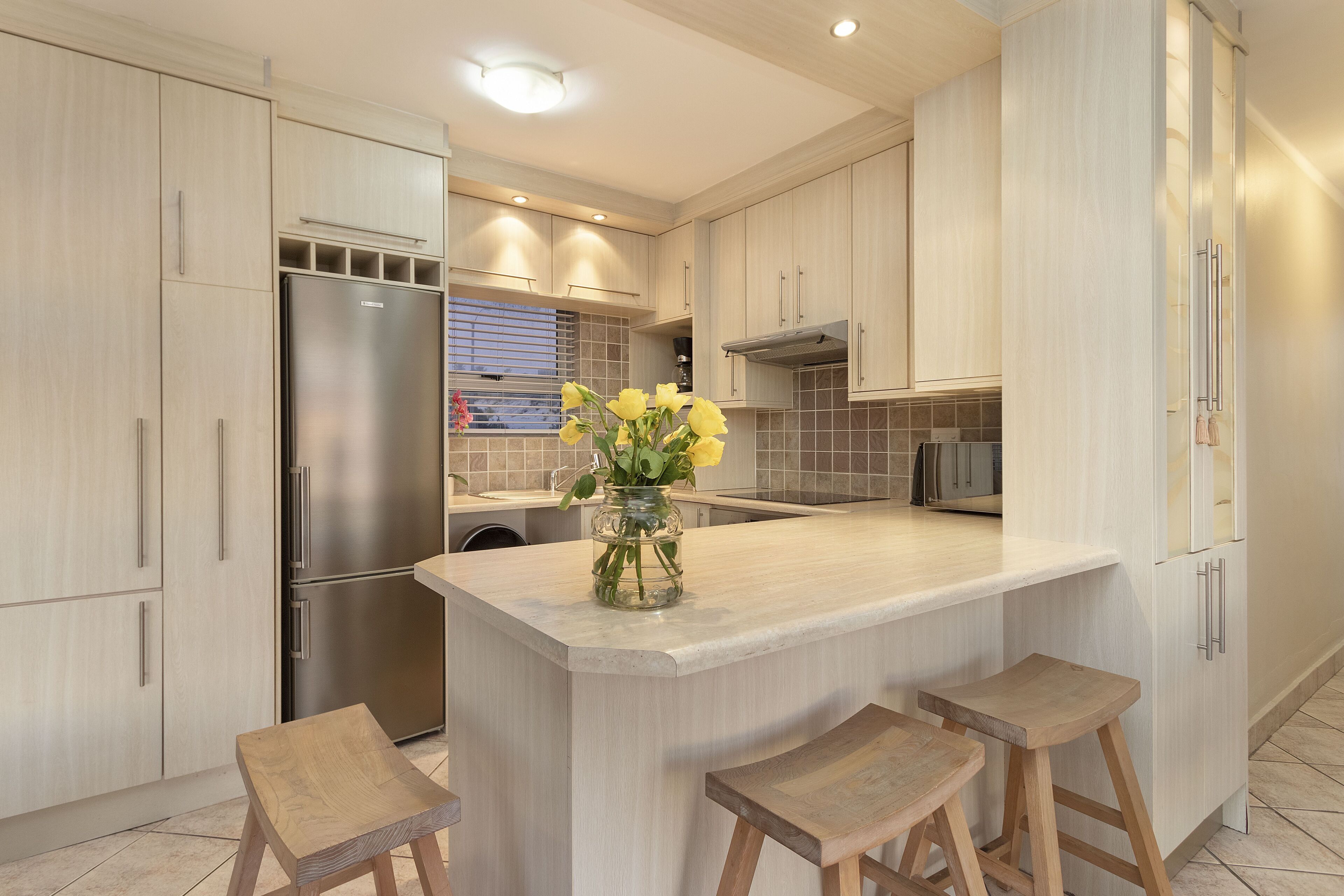 A modern kitchen with light wood cabinets, a stainless steel refrigerator, a central island with stools, and a vase of yellow flowers.