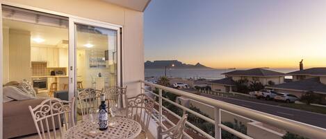 A balcony with a table and chairs, overlooking a street and a body of water during sunset.