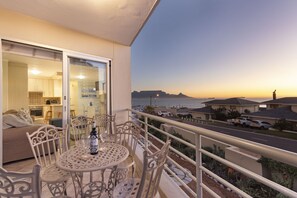 A balcony with a table and chairs, overlooking a street and a body of water during sunset. - The Bay A101 (Cape Town)