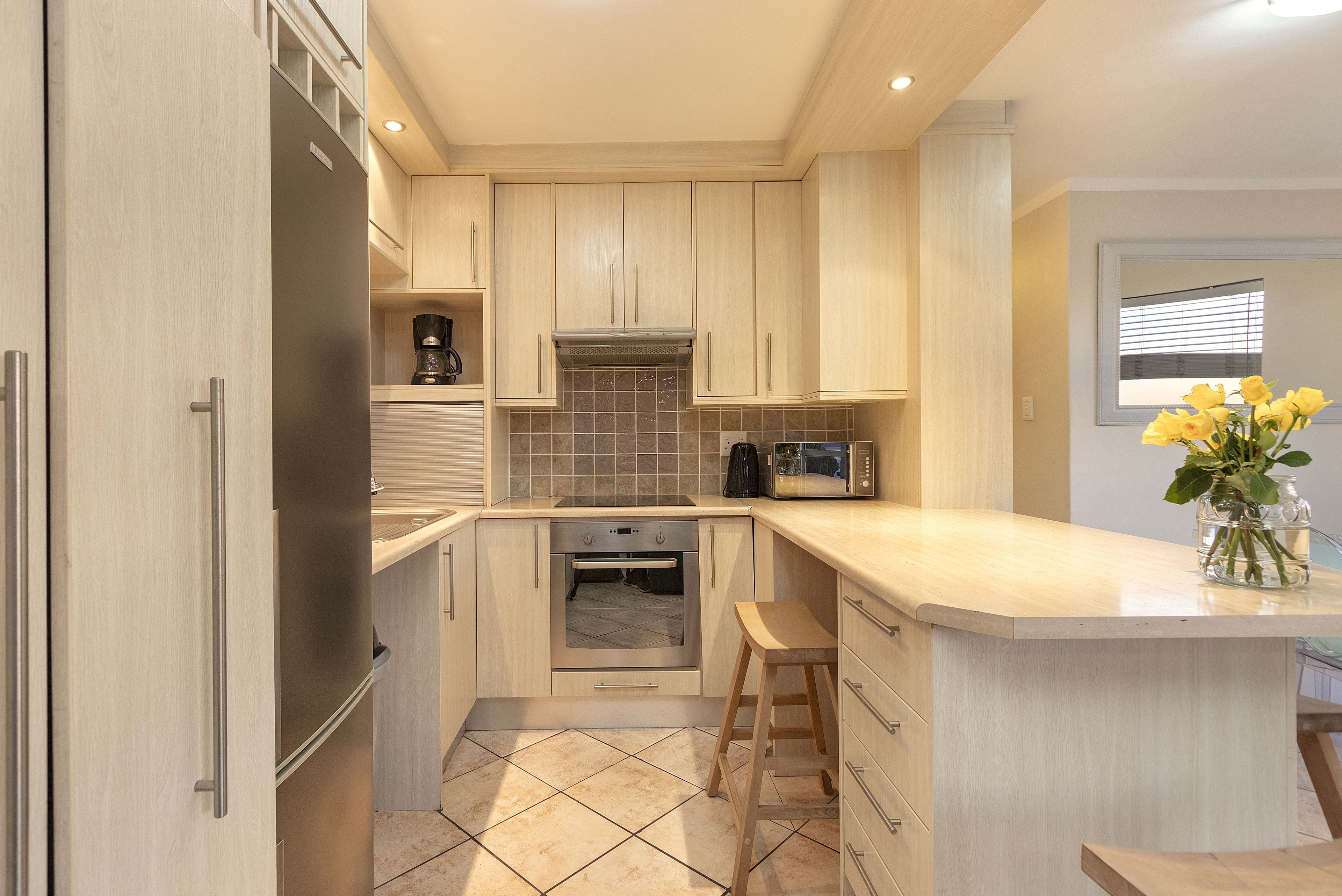 A modern kitchen with light wood cabinets, a stainless steel refrigerator, a built-in oven, and a microwave. There is a small stool and a vase with flowers on the counter.