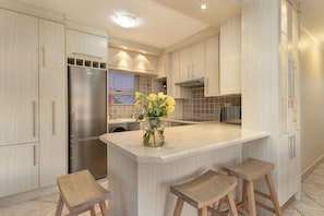 Une cuisine moderne avec des meubles de couleur bois clair, un réfrigérateur en acier inoxydable, un îlot central avec des tabourets et un vase rempli de fleurs jaunes.