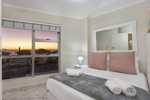 A hotel room with a large bed, a nightstand with a lamp, a window with a view of a sunset, and a mirror above the bed. - The Bay A101 (Cape Town)