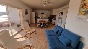 A living room with a blue sofa, wooden chairs, a dining table with chairs, a kitchen area, and a ceiling fan. - Villa close to the beach with exceptional views. (Port-la-Nouvelle)