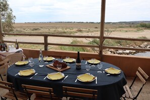 Mesa lista para una comida con botella de vino, copas y platos, con vista a un paisaje natural.