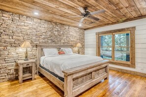 Een slaapkamer met een stenen muur, houten plafond, een bed met witte beddengoed, twee nachtkastjes met lampen, en een groot raam met uitzicht.