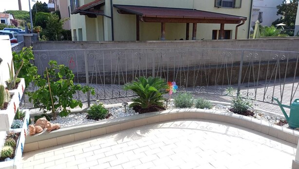 A paved courtyard with a curved bench, various plants, and a metal fence. - Newly furnished two-room apartment, 800 mt from the sea. (Cecina)