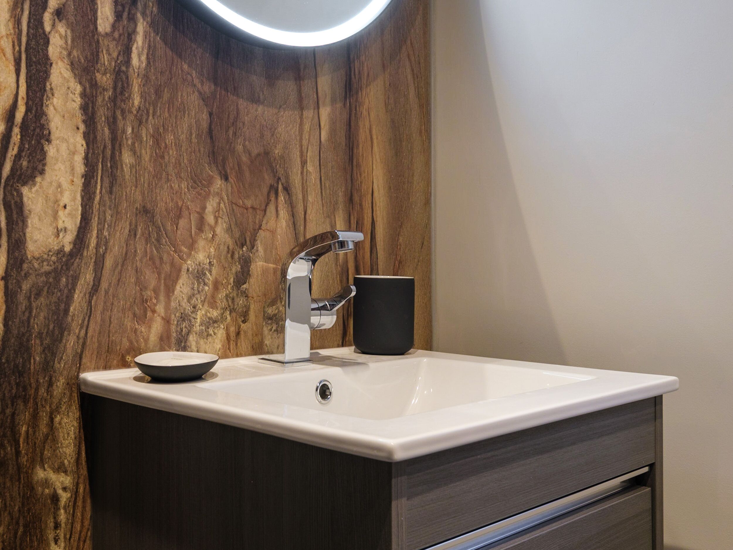 A modern bathroom with a marble wall, a white sink, a chrome faucet, and a black mug.