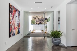 A modern hallway with a large abstract painting, a potted plant, and a view into a room with a tiled floor and a rock garden. - Stay in 3rd Rock Home ! (Palm Springs)