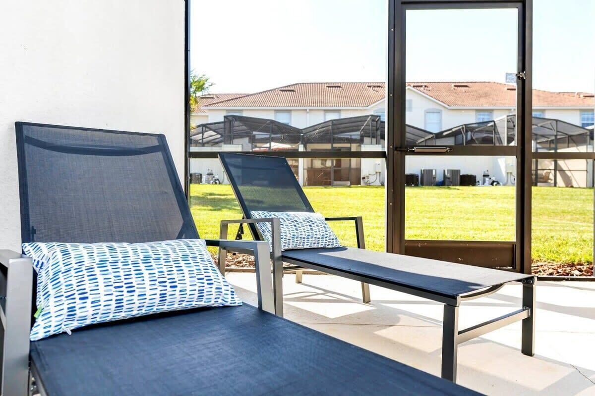 A sunlit outdoor lounge area with two reclining chairs and a bench, featuring a clear view of a residential neighborhood through large glass doors.