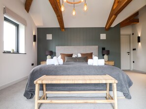 A modern bedroom with a large bed, wooden beams, a bench, bedside lamps, and a window with a plant.