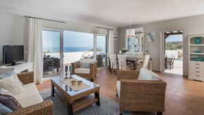 A spacious living area with a large window overlooking the sea, wicker furniture, a dining table, and a television. - Villa Melodie, fantastic pool & terraces, sea view (Vamos)