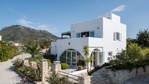 A modern white building with a stone pathway leading to the entrance, surrounded by greenery and a mountainous backdrop. - Villa Melodie, fantastic pool & terraces, sea view (Vamos)