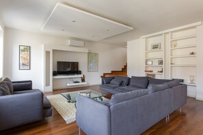 A modern living room with a grey sectional sofa, a glass coffee table, and a flat-screen TV. There is a staircase leading to an upper level, and a white shelving unit with decorative items. - Point of View (Verona)