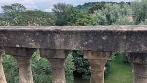 Une balustrade en pierre, ornée de colonnes, surplombe un parc verdoyant et luxuriant, parsemé d’arbres, et offre une vue sur une colline lointaine.