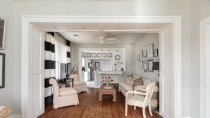 A spacious living room with a wooden floor, a coffee table, and a sofa. There is a kitchen area in the background with a refrigerator and a clock.