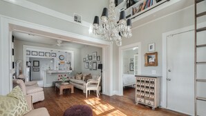 A spacious living area with a wooden floor, a chandelier, and a kitchenette in the background.