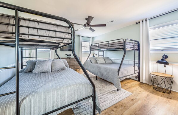 A bunk bed room with a ceiling fan, a small table with a lamp, and a window with blinds.