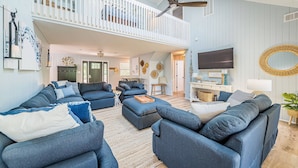 A spacious living room with a high ceiling, a balcony, and a large TV. - Beach Blessing - Southern Shores, 225 to Beach Access, Quiet Area, Hot Tub, Pets OK, Flex Stays (Southern Shores)