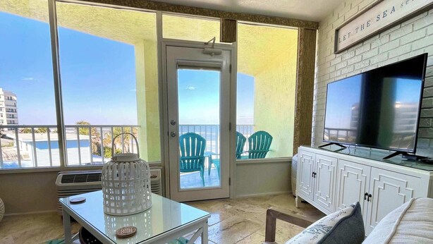 Een balkon met uitzicht op zee, een televisie, een witte kast en een vloerkleed met bloemenprint.