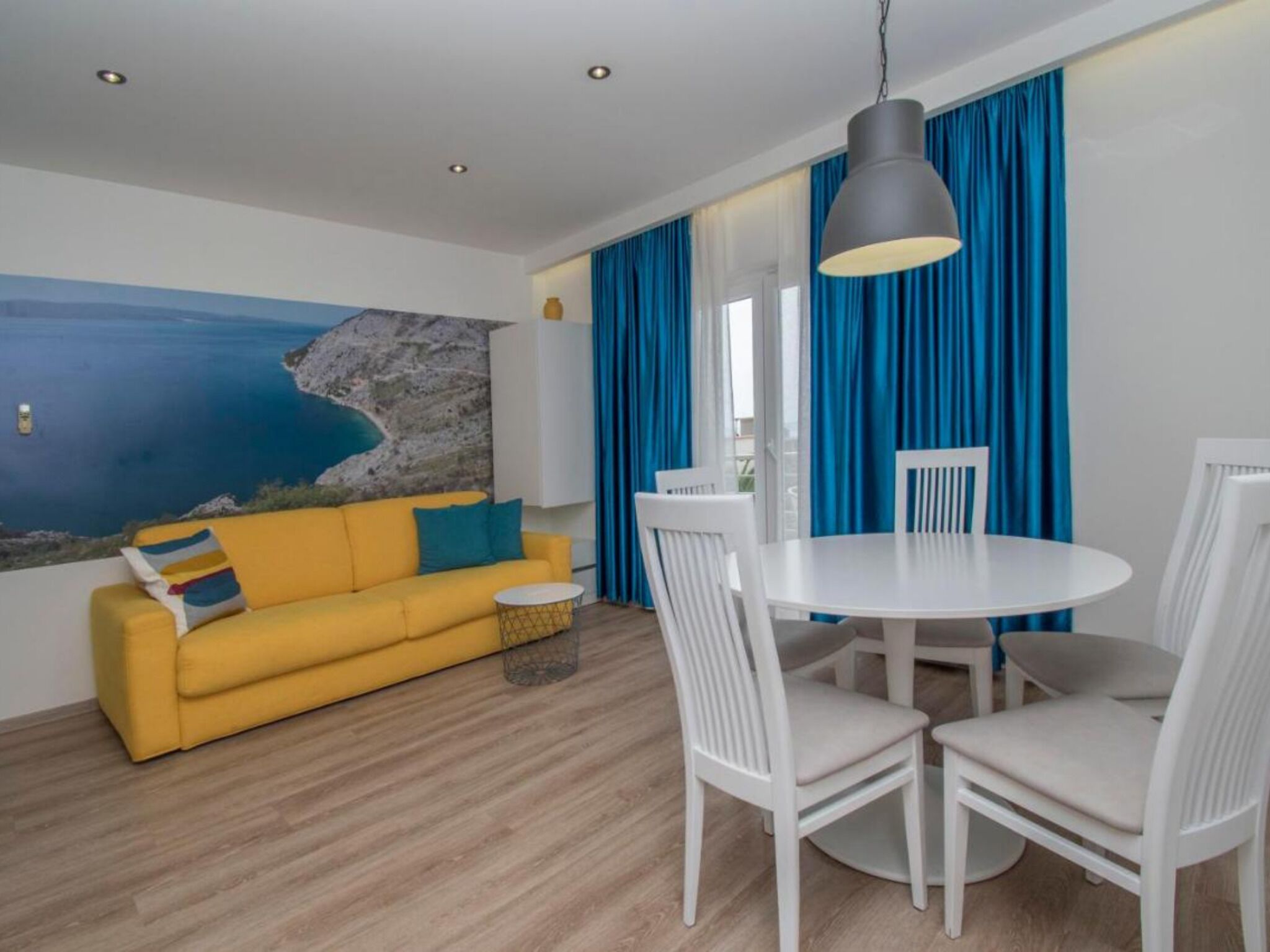 Una habitación con un sofá amarillo, una mesa de comedor blanca y sillas, un ventanal con cortinas azules y un cuadro con una escena costera en la pared.