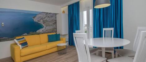 Una habitación con un sofá amarillo, una mesa de comedor blanca y sillas, un ventanal con cortinas azules y un cuadro con una escena costera en la pared.