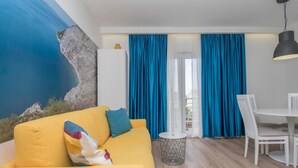 A modern living room with a yellow sofa, a dining table with white chairs, and a view of the sea through large windows with blue curtains. - Apartments Antonio (Tucepi)