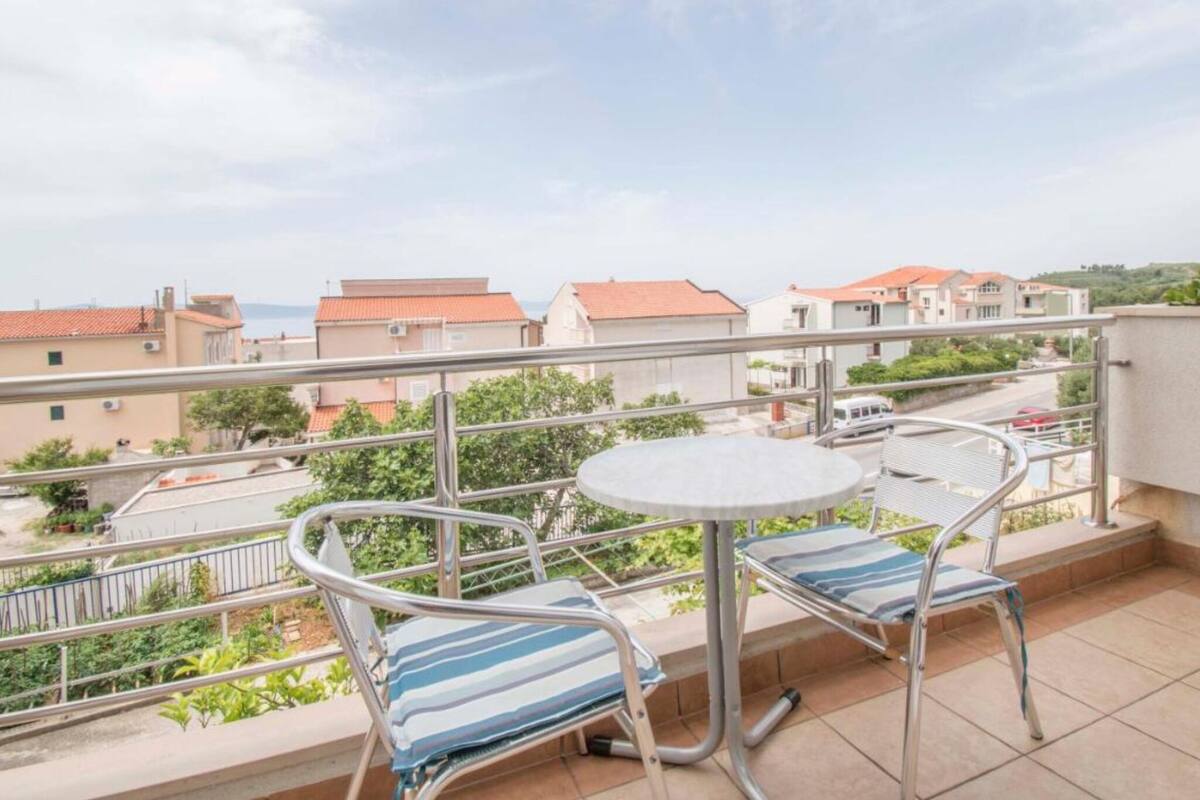 A balcony with a round table and two striped lounge chairs, overlooking a residential area with houses and greenery.